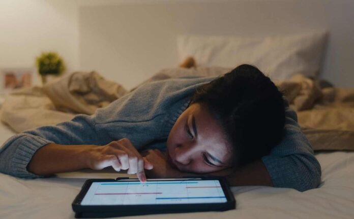 A female laying face down on a bed scrolling on her laptop. How Blue Light Affects Sleep.