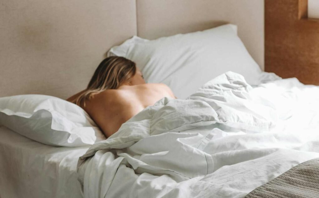 A woman laying face down on a bed with white bedding. Her naked upper back is visible. A good night's sleep starts with choosing the right mattress.