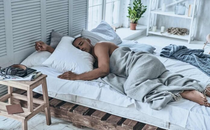 A male asleep on a white lined sheet with a grey blanket over him.