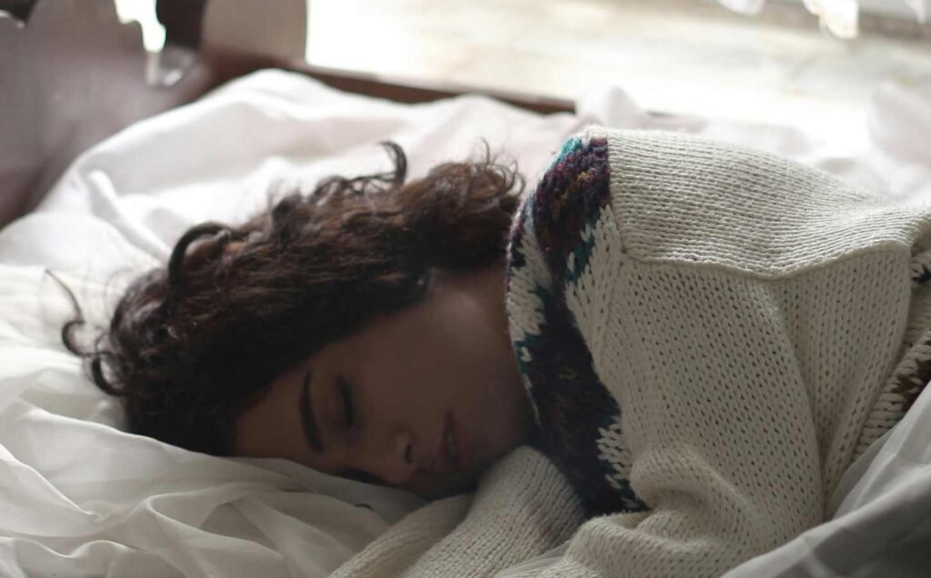 Head and shoulders of a female asleep on white sheets. She is wearing a woolen cardigan.