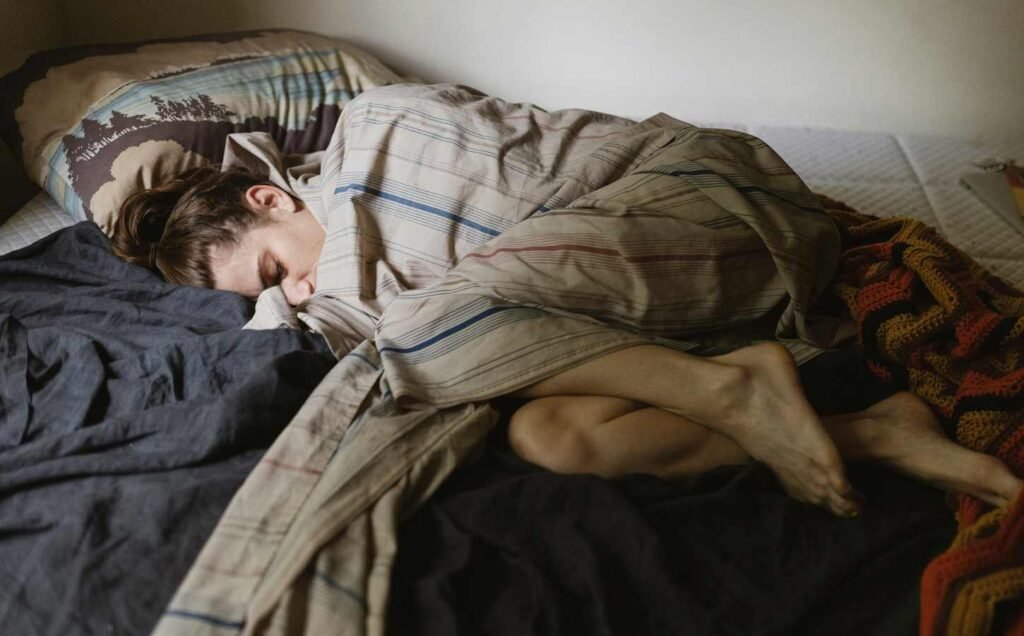 A young woman asleep in the fetal position. Her lower legs can be seen under a multi colored sheet that covers the rest of her body.