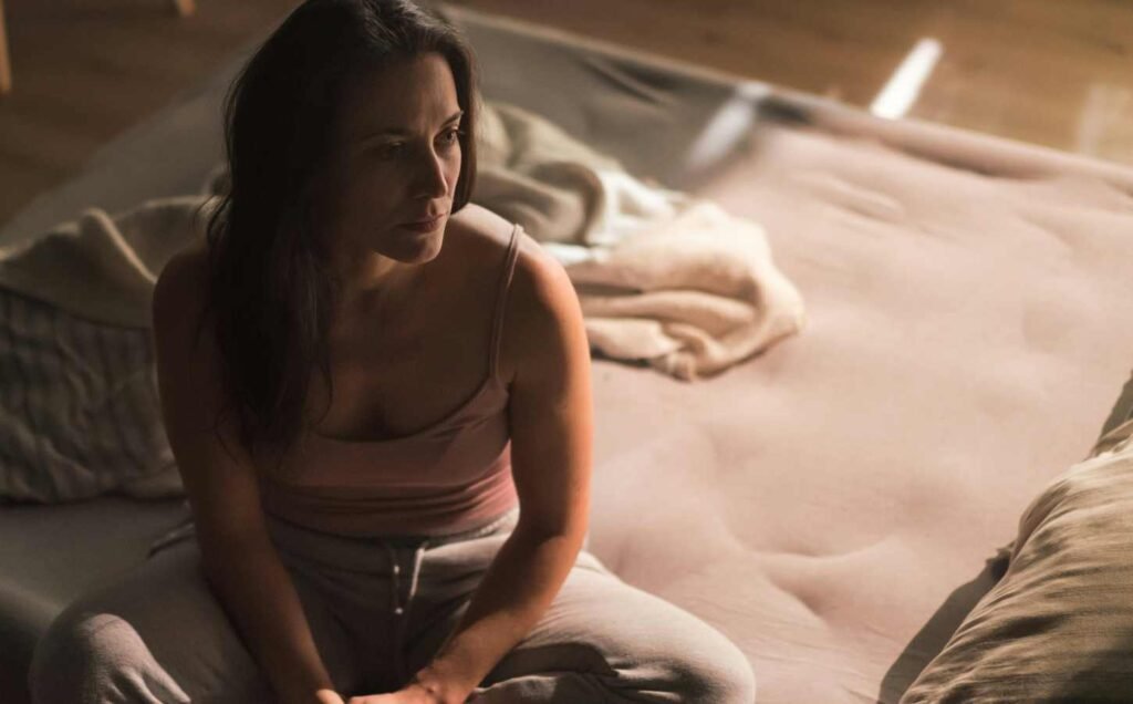 A woman in a pink singlet and grey track pants sitting on the edge of a bed, unable to sleep.