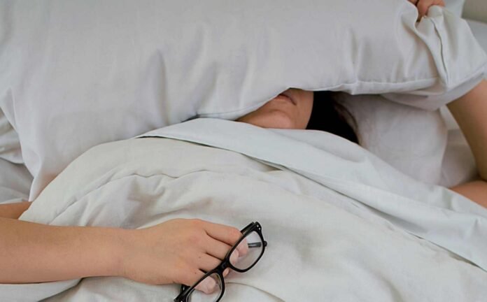 A woman hiding under her pillow, holding her reading glasses in her right hand. Related to the question, can restless leg syndrome intensify.