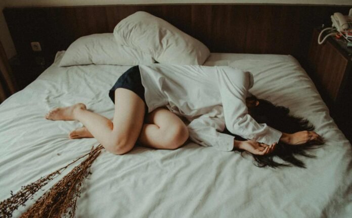 Female laying sideways on a bed, clothed in a white shirt and black shorts. Illustrating the disruptive power of restless leg syndrome.