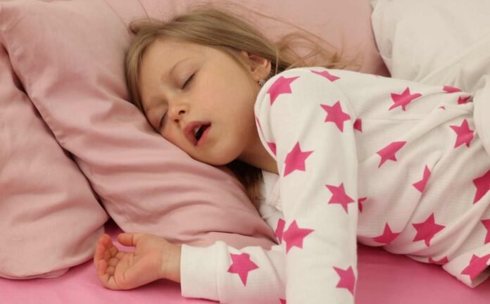 A little girl in bed wearing pink and white pajamas snoring.