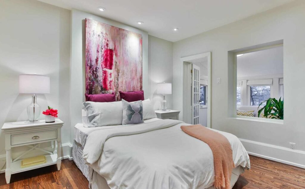 A luxury bedroom, mostly white, cream and pastel colors. With an original artwork hung above the bedhead.