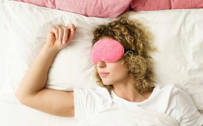 A blonde woman sleeping with a pink blindfold. Wearing a white night shirt on white sheets.