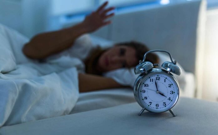 An alarm clock a few minutes before 4:00 am, with a woman gesturing with her hand in the background.