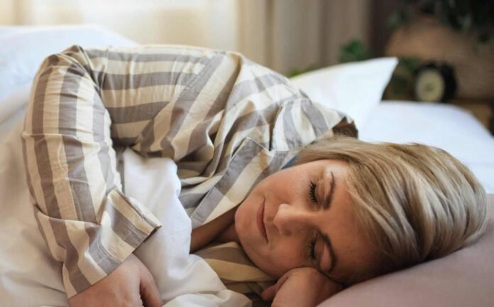 A woman asleep in a bed with white sheets and pink pillow. She is wearing white and light brown striped night shirt.