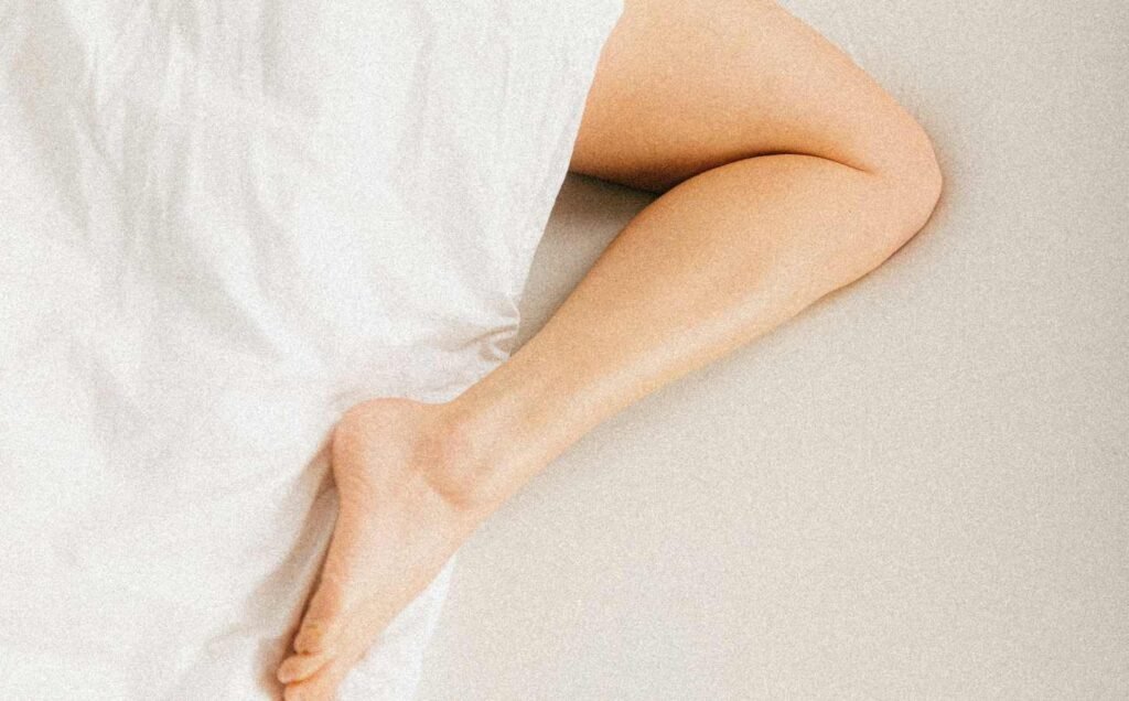 The lower torso of a woman sleeping beneath a white sheet with her lower leg exposed. The complexity of restless leg syndrome.