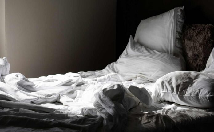 An unmade bed, ruffled white bedding against a brown wall.