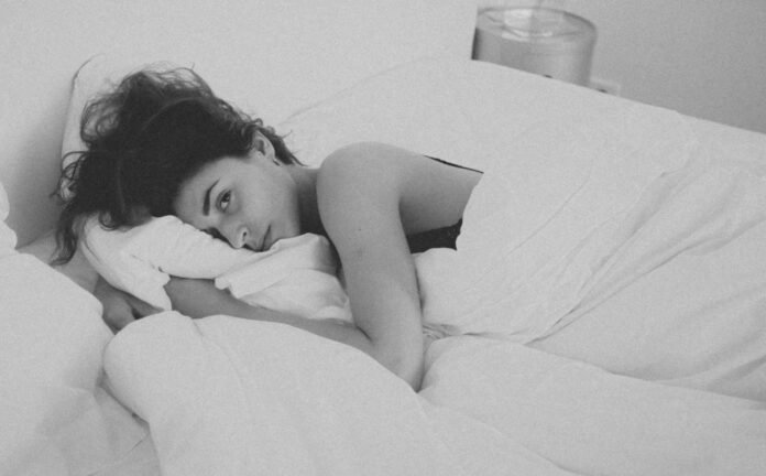 A black and white photo of a dark haired female laying awake in bed, looking at the camera.