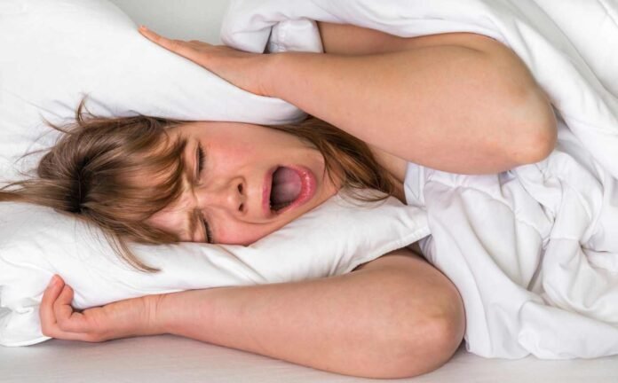 A woman who can't sleep, yawning and wrapping a pillow around her ears.