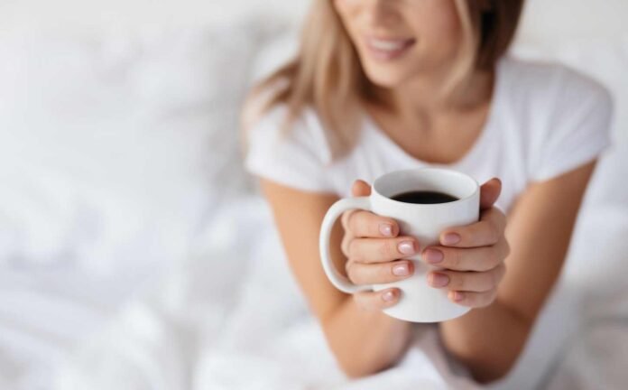 A woman in a white t-shirt sitting up in bed drinking coffee.