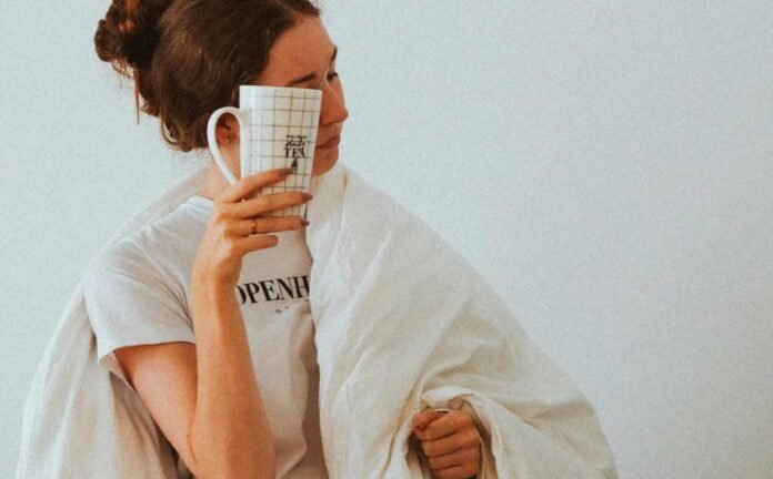 a woman holding a mug up to her face, holding a blanket over her shoulder, looking very tired during the daytime.