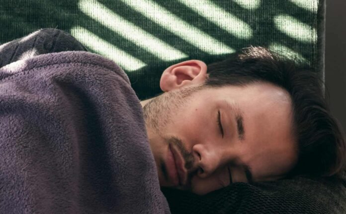 A male laying down on a green couch asleep with a purple blanket up to his shoulders.