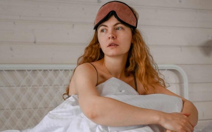 A woman sitting up in bed with an eye mask pushed up over her forehead.