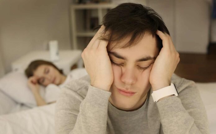 A male sitting on the edge of a bed, head in hands, unable to sleep. A female sleeps in the background.