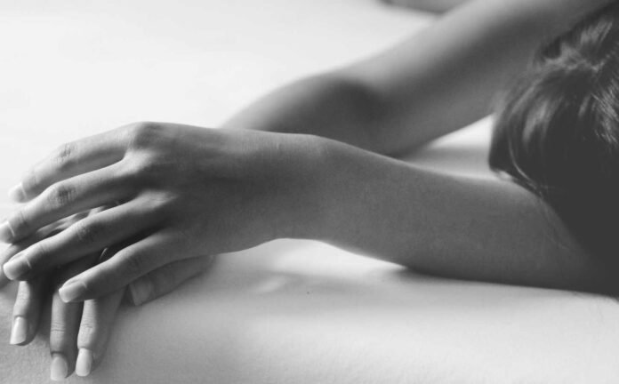 Close up, black and white photo, of a female laying on a bed, arms s[played above her head.