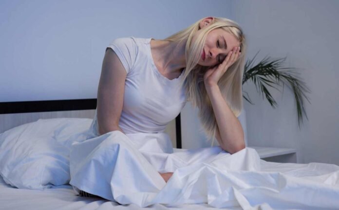 A blobe woman sitting up in bed, head resting in one hand, possibly experiencing irregular sleep wake rhythm disorder.