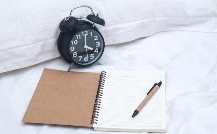 A black alarm clock, a pencil and a diary on a bed with white cover and pillow case. This person is keeping a sleep diary to help with a sleep disorder.