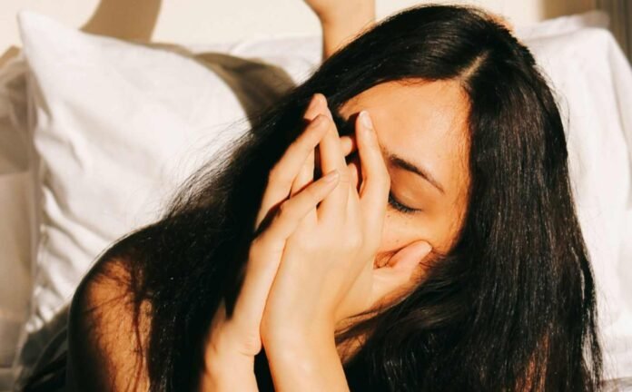 Close up of a dark haired woman laying on her bed with both hands cupped against her face.