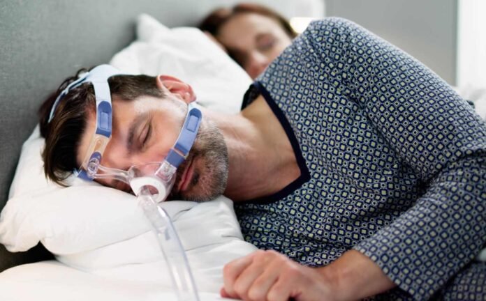 A male and female in bed asleep. The male is wearing a CPAP machine.