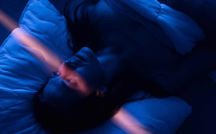 A woman laying in bed with a streak of light across her face in an otherwise bark blue background. Indicative of undergoing the Multiple Sleep Latency Test