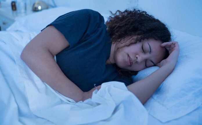 A black female trying to sleep. Wearing a blue t-shirt, on light blue sheets.