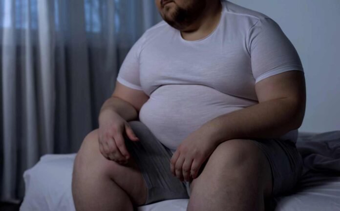 An obese male sitting on the edge of a bed, possibly experiencing obesity hypoventilation syndrome.