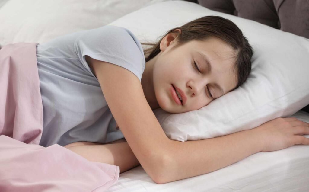 A young girl asleep in bed, one arm raised to one side.