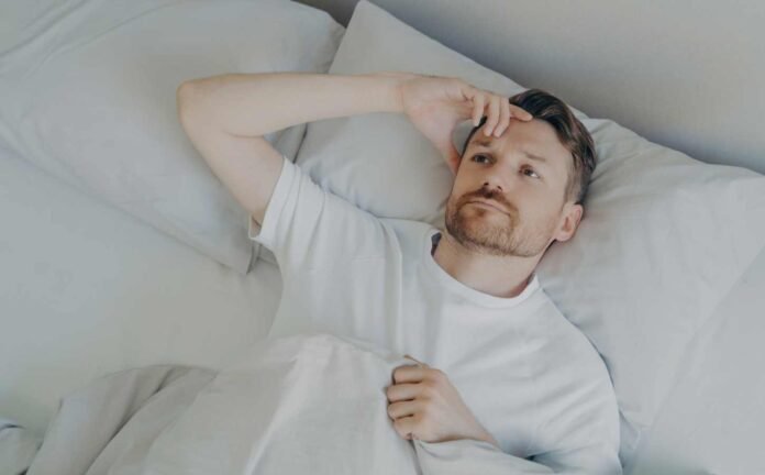 Man laying in bed unable to sleep. Has one hand raised to his forehead.