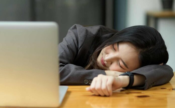 An Asian woman who has fallen asleep at her desk - a sleep attacks. She is slumped on her wooden deck in front of her laptop computer.