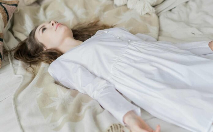 A female in a white nightdress laying on a bed experiencing sleep paralysis.