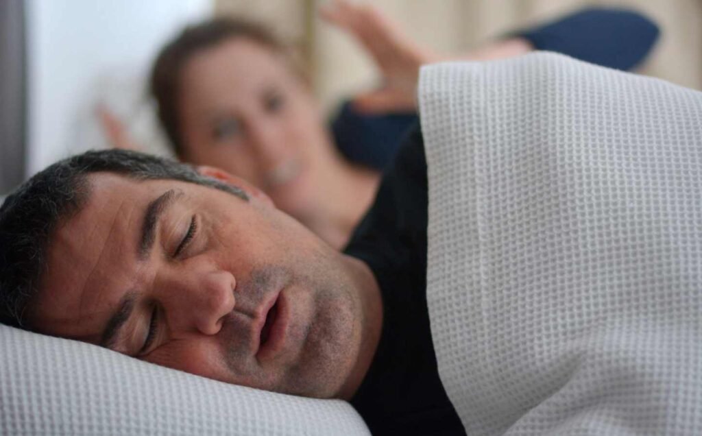 A male and female in bed. The male is asleep snoring, the female in the background expressing her frustration at the noisy snoring.