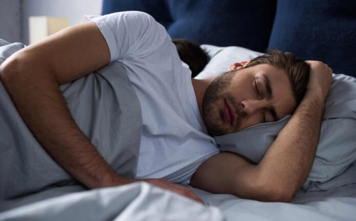 A male in a white t-shirt asleep in bed, one arm above his head.