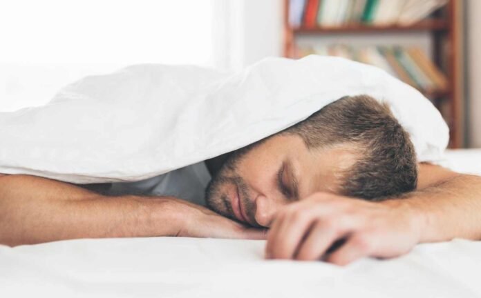 A man laying face down in bed asleep, with a white sheet pulled up over his head.