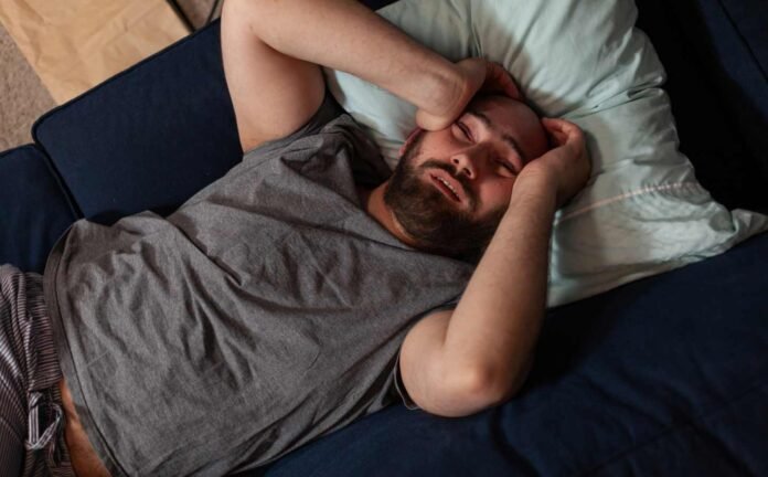 A man in bed on his back, hands rubbing his forehead as he is unable to sleep, caught up in the wake state of the sleep-wake-cycle.