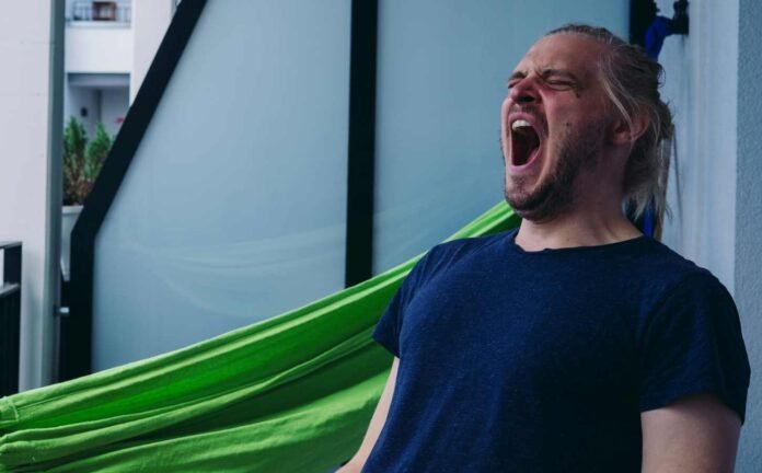 A man on a balcony, with a bright green hammock behind him. He is having a massive yawn.