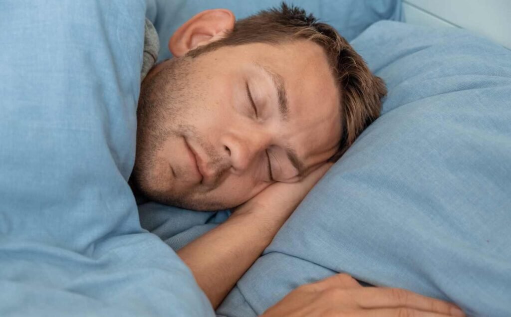 Head and arms of a man in bed sleeping on blue sheets and pillow.