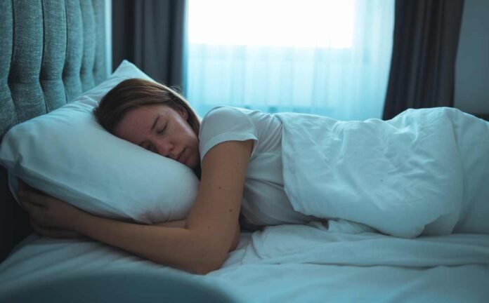 A female in a white t-shirt sleeping on her stomach in bed, the room has a light blue tinge.