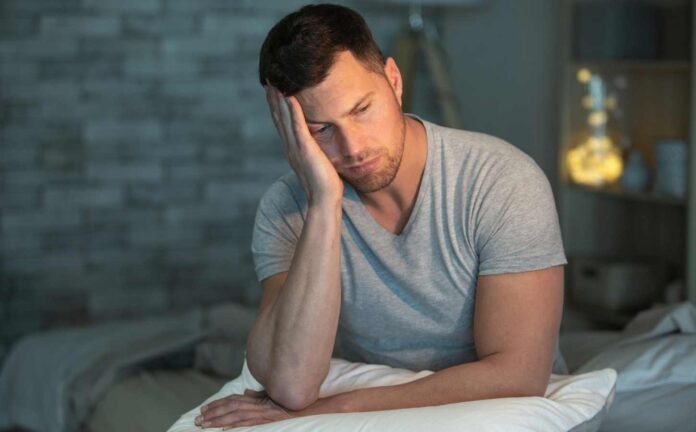 A man in a grey t-shirt sitting up in bed with insomnia.