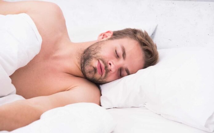 A man asleep, in a bed with white sheets. He is on his side facing the camera.