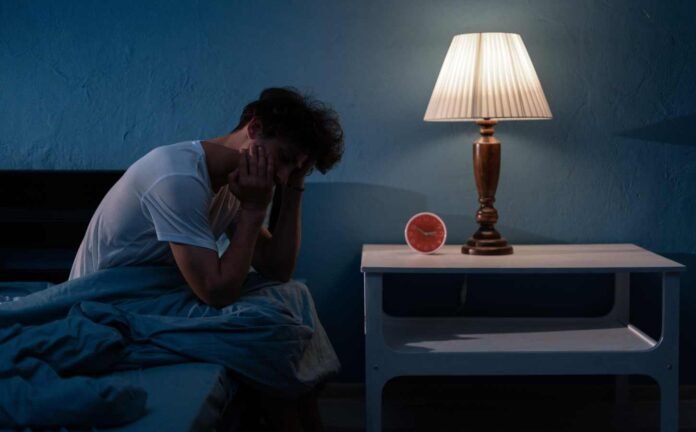 A man siting on the side of his bed at 2:50 am, he's tired but cant sleep.