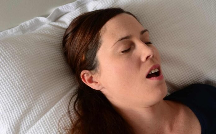 A woman in a black t-shirt in a bed with white sheets and pillow, asleep and snoring.