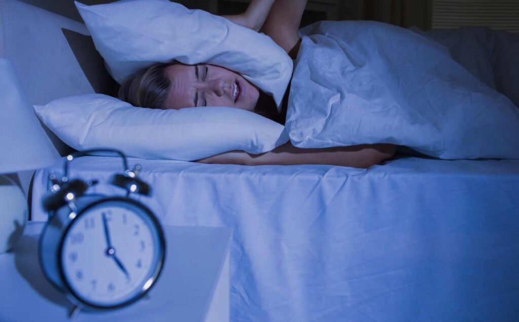 A woman putting pillows over her ears with an alarm clock in the foreground showing it's 5 AM.