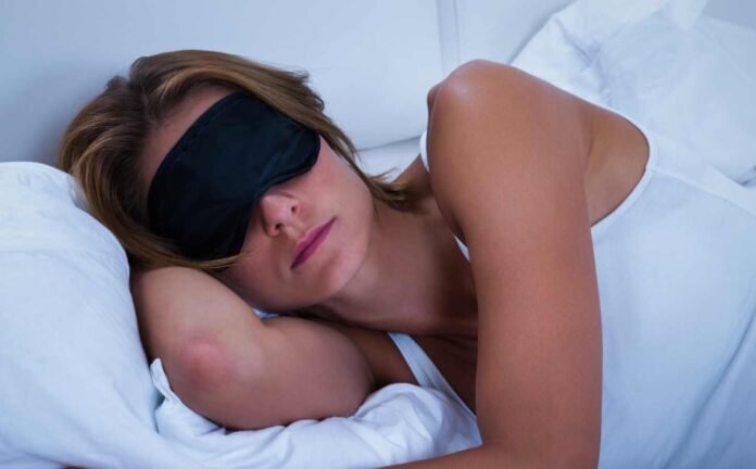 Woman in a white singlet, on white bedding, asleep with a black sleep mask.