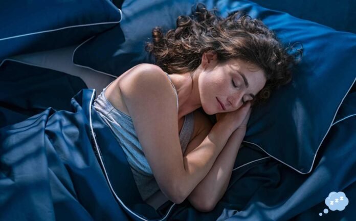 A woman asleep in a bed with dark blue sheets wearing a light blue might dress.