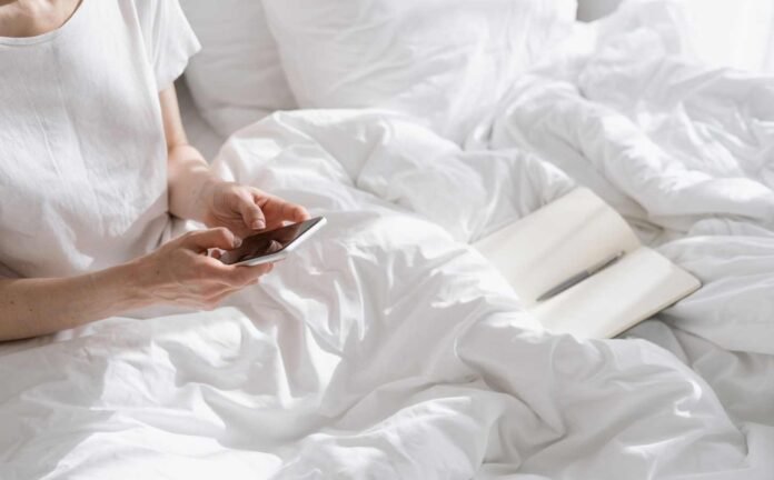 A woman sitting in bed holding her smart phone surfing the internet.