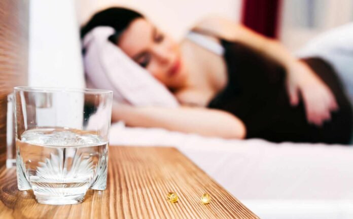 A half full glass of water on a night stand beside a bed. In the background is a woman in a black singlet asleep on a bed.